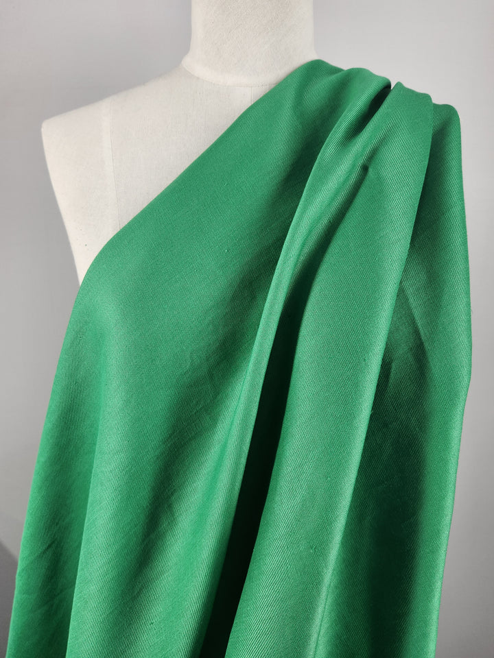 A mannequin is elegantly draped with Super Cheap Fabrics' Pure Washed Linen in Jelly Bean, 140cm wide. This green, lightweight linen is soft with a slight texture, forming gentle folds and shadows against a plain gray background.