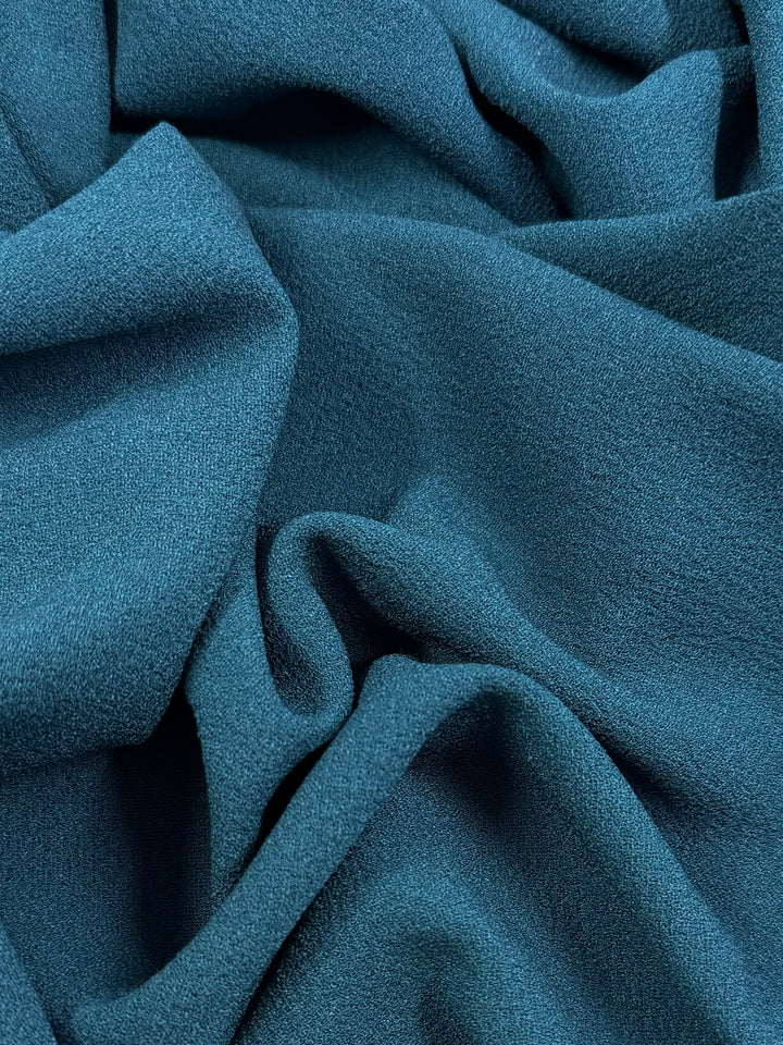 A close-up of Super Cheap Fabrics' Plain Crepe - Teal - 150cm shows soft folds and wrinkles, highlighting its smooth yet slightly grainy medium-weight polyester texture.