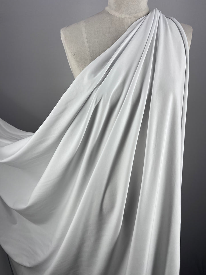 A white 180cm Cotton Lycra fabric by Super Cheap Fabrics drapes elegantly over a dress form on a gray background. Its stretchy nature allows for smooth folds and fluid movement, reminiscent of bodycon dresses.