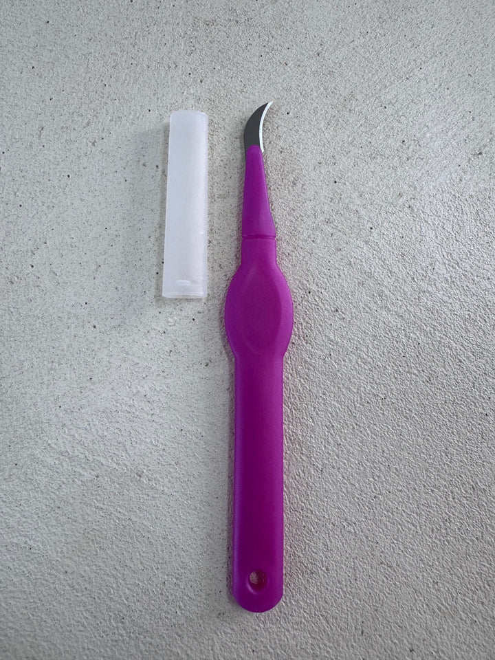 A pink seam ripper by Super Cheap Fabrics rests on a textured gray surface, featuring a precision curved blade and a long white cap.