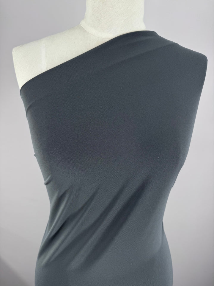 A white mannequin showcases a dark gray, one-shoulder garment made from Super Cheap Fabrics’ ITY Knit - Charcoal (150cm), draped diagonally across the torso and displayed against a plain light background.
