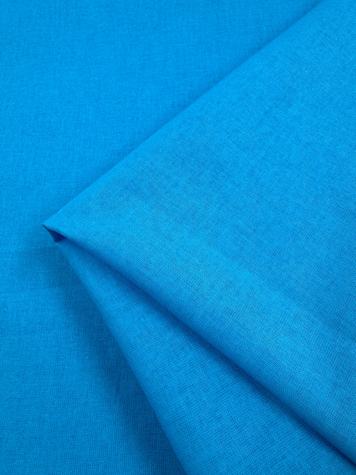 A folded piece of Super Cheap Fabrics' Plain Cotton - Bachelor Button - 112cm, featuring a slightly textured surface, rests on a matching blue background.