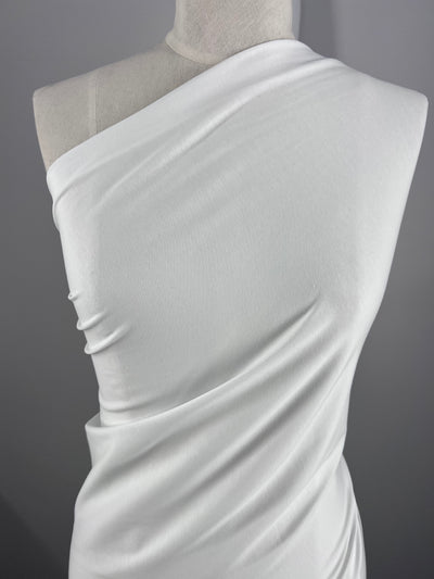 The Cotton Lycra - White 180cm fabric by Super Cheap Fabrics is elegantly draped on a mannequin, displaying its smooth, stretchy material with subtle folds and contours against a neutral gray background.
