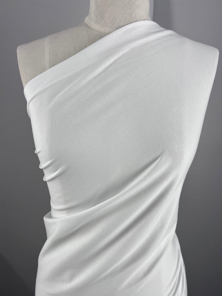 The Cotton Lycra - White 180cm fabric by Super Cheap Fabrics is elegantly draped on a mannequin, displaying its smooth, stretchy material with subtle folds and contours against a neutral gray background.