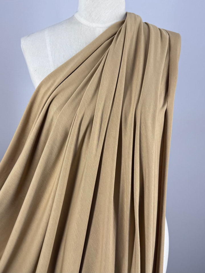 A beige Cotton Lycra fabric from Super Cheap Fabrics, draped over a white mannequin with soft folds, creates a flowing effect. The smooth-textured Doe 160cm material covers one shoulder and cascades down the front against a plain light gray background.