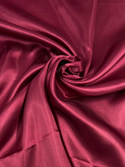 Super Cheap Fabrics’ Satin - Red Bud - 150cm features deep red satin with gentle folds and a spiral pattern that reflects light, highlighting its smooth, shiny, and luxurious texture—perfect for adding elegance to any design.