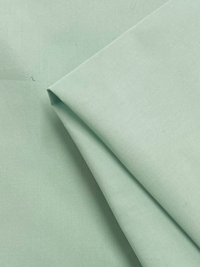 A close-up of Super Cheap Fabrics’ Plain Cotton - Aqua Foam - 150cm shows its neatly folded, soft light green fabric with a smooth finish. Made from 100% cotton, this lightweight fabric combines comfort and elegance.