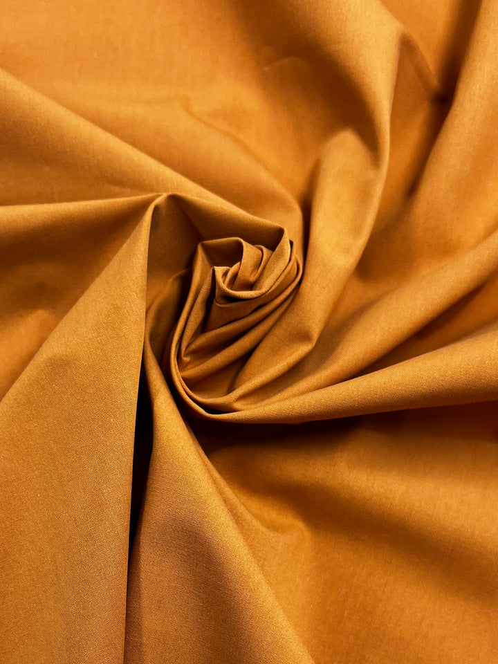 A close-up of Super Cheap Fabrics' Plain Cotton - Marmalade - 150cm displays its rust-orange color, lightweight feel, and smooth, soft folds arranged in a spiral.