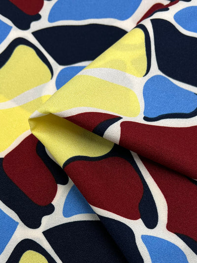A close-up of Super Cheap Fabrics' Printed Crepe - Bovine Rainbow (150cm) shows abstract shapes in red, yellow, blue, white, and black—ideal for lightweight high-fashion designs with its modern pattern and vibrant polyester finish.