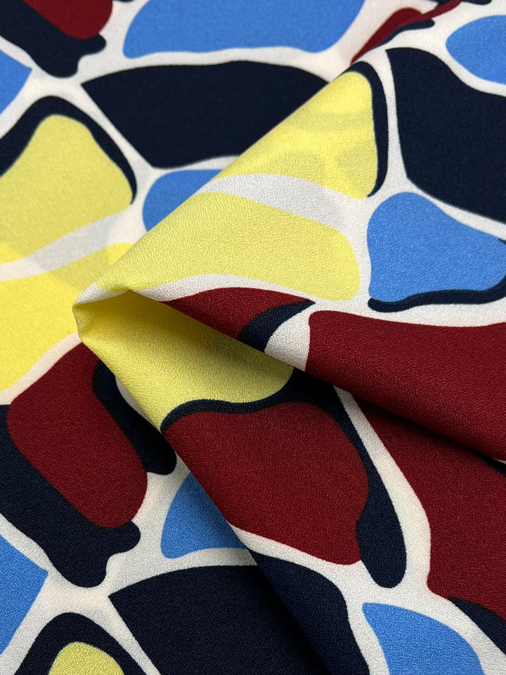A close-up of Super Cheap Fabrics' Printed Crepe - Bovine Rainbow (150cm) shows abstract shapes in red, yellow, blue, white, and black—ideal for lightweight high-fashion designs with its modern pattern and vibrant polyester finish.