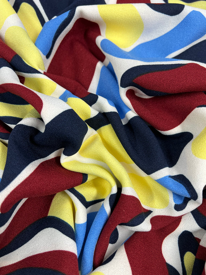 A close-up of Super Cheap Fabrics' Printed Crepe - Bovine Rainbow (150cm) shows vibrant crumpled polyester with an abstract red, yellow, blue, black, and white pattern for a dynamic visual texture.