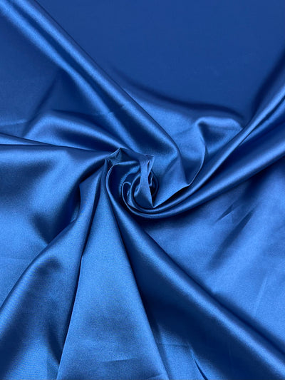 A close-up image of a smooth and shiny blue Super Cheap Fabrics Satin Deluxe - Royal - 150cm. The light-weight material is slightly wrinkled and gathered in the center, creating soft folds and textures that highlight its lustrous and reflective quality.