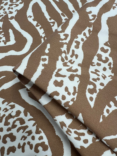 A close-up view of folded fabric showcasing the Printed Bengaline - Jungle Fusion by Super Cheap Fabrics, featuring an abstract animal print pattern in shades of light brown and white. This medium-weight fabric, ideal for skirts, dresses, or pants, is intricately woven with a blend of organic and geometric shapes reminiscent of zebra or cheetah prints. The fabric measures 150cm in width.