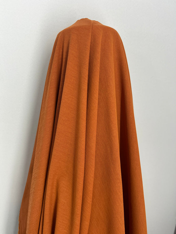 Against a light grey background, an individual is wrapped in Super Cheap Fabrics' Gold Flame Suiting (144cm), featuring a light to medium weight orange polyester fabric, resembling a ghostly figure with hints of shimmering gold flame.
