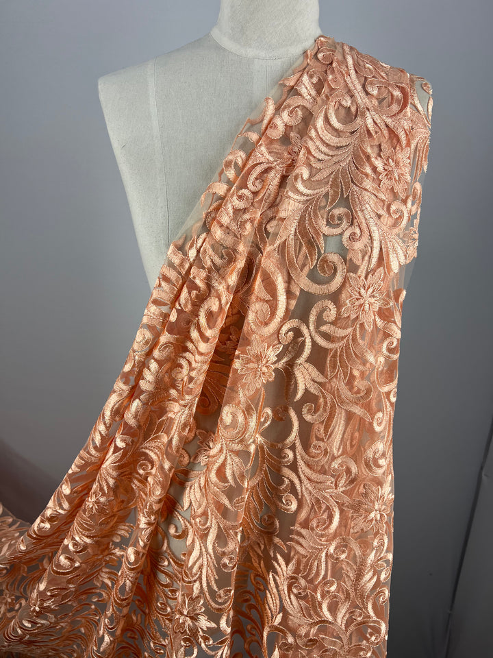 A dress form draped with sheer peach lace fabric featuring intricate floral and swirl embroidery. The Evening Lace - Galadriel from Super Cheap Fabrics hangs elegantly, showcasing its delicate and ornate design.