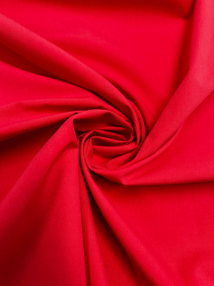 A close-up of Super Cheap Fabrics’ Plain Cotton - Watermelon (150cm), lightweight and fiery red, twisted into a spiral to reveal smooth, flowing folds and creases.