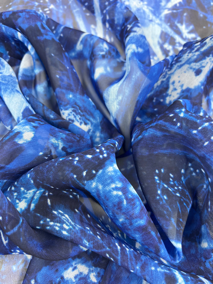 A close-up of Super Cheap Fabrics' Printed Chiffon - Galleria (150cm) reveals a vibrant blue and white abstract pattern. The silky, translucent material showcases dynamic folds and textures that evoke high-fashion elegance.