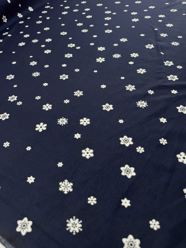The Super Cheap Fabrics Printed Cotton Lycra - Twinkle (160cm) is a Navy fabric with small white snowflake patterns, crafted from Cotton Spandex for a wintry and festive look.