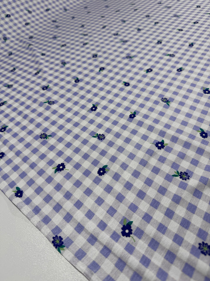 The Printed Cotton Lycra - Lavender Ditsy from Super Cheap Fabrics features a soft lavender and white checkered pattern, adorned with small blue flowers on a 153cm-wide cotton spandex fabric that lies flat to showcase its delicate design.