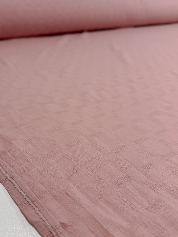 A close-up of Super Cheap Fabrics’ Textured Cotton - Misty Rose (140cm), a lightweight, 100% cotton fabric in light pink with a subtle textured square pattern, partially unrolled on a flat surface.