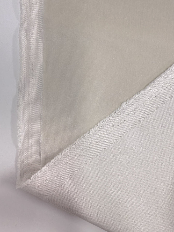 A close-up of Super Cheap Fabrics' Oxford Velvet - Warm Snow (144cm) shows the heavyweight, slightly sheer off-white cotton with a frayed selvage and smooth texture, folded at the corner.