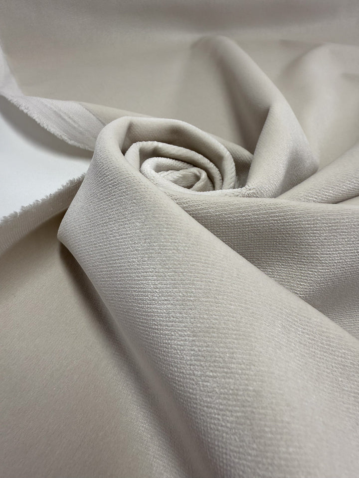 A close-up of Oxford Velvet - Warm Snow - 144cm by Super Cheap Fabrics, shows soft beige heavyweight cotton velvet with gently swirling folds and a visible selvage edge; the smooth texture has a slightly matte finish.