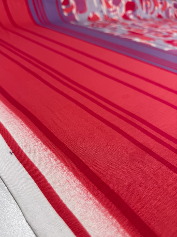 Close-up of Super Cheap Fabrics’ Viscose Knit - Abstract Striped Geo (150cm), featuring smooth, stretchy viscose fabric with diagonal red and pink stripes, a white edge, and a partially visible background pattern.