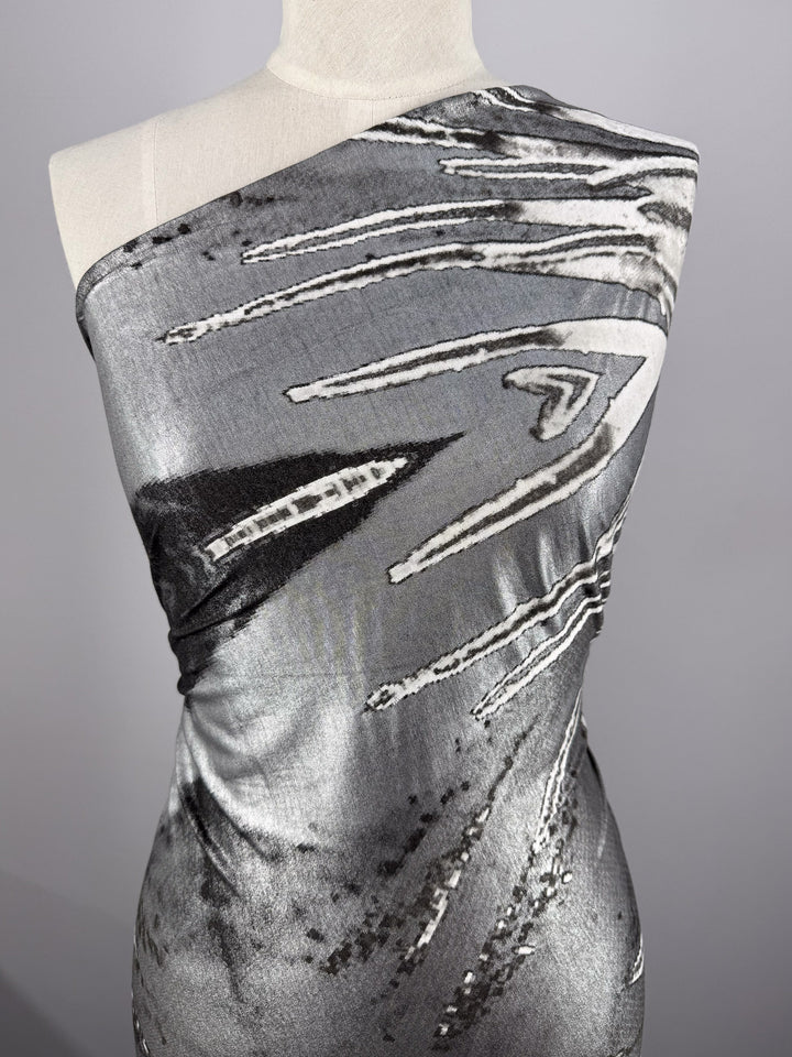 A headless dress form draped in Super Cheap Fabrics' Viscose Knit - Cosmic Burst (145cm), featuring a shiny, one-shoulder design with an abstract black, white, and gray bold-lined pattern.