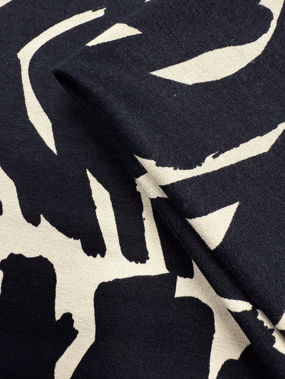 Close-up of Super Cheap Fabrics' Viscose Knit - Tribal Brushstroke (142cm): a lightweight black stretch fabric featuring cream abstract brushstroke patterns, with a fold highlighting its texture and thickness.