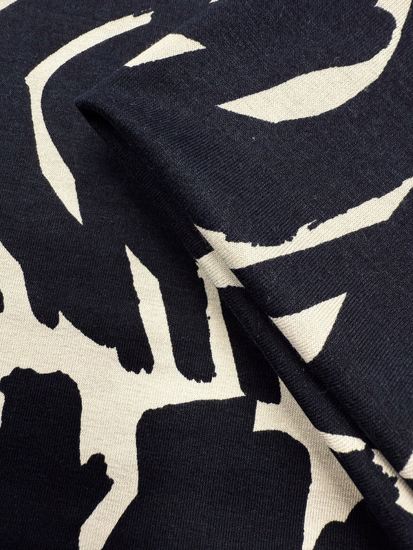 Close-up of Super Cheap Fabrics' Viscose Knit - Tribal Brushstroke (142cm): a lightweight black stretch fabric featuring cream abstract brushstroke patterns, with a fold highlighting its texture and thickness.