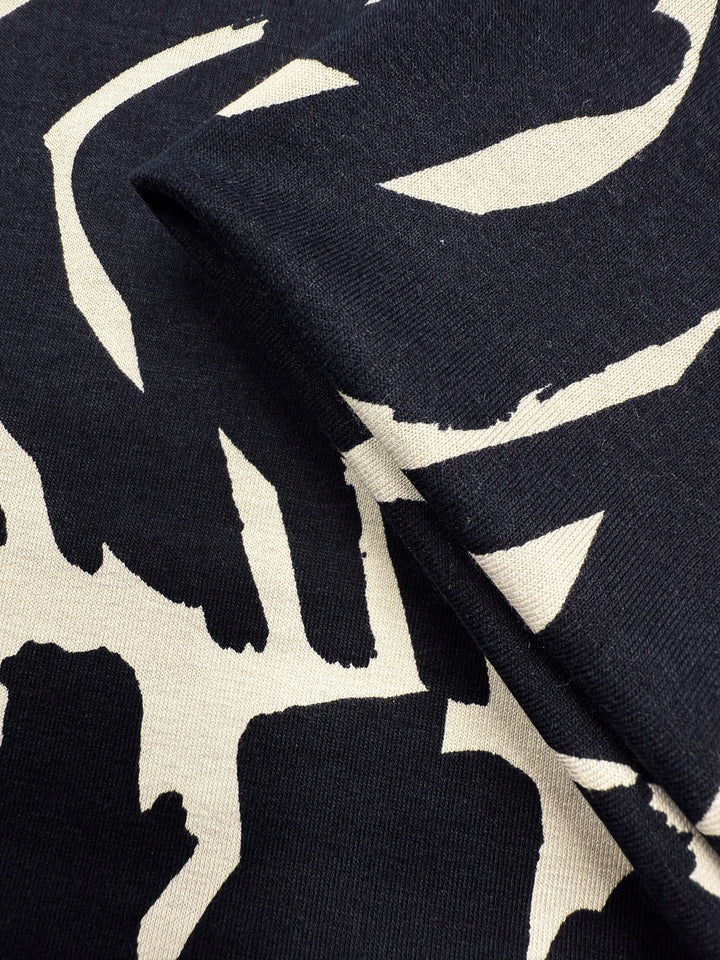 Close-up of Super Cheap Fabrics' Viscose Knit - Tribal Brushstroke (142cm): a lightweight black stretch fabric featuring cream abstract brushstroke patterns, with a fold highlighting its texture and thickness.