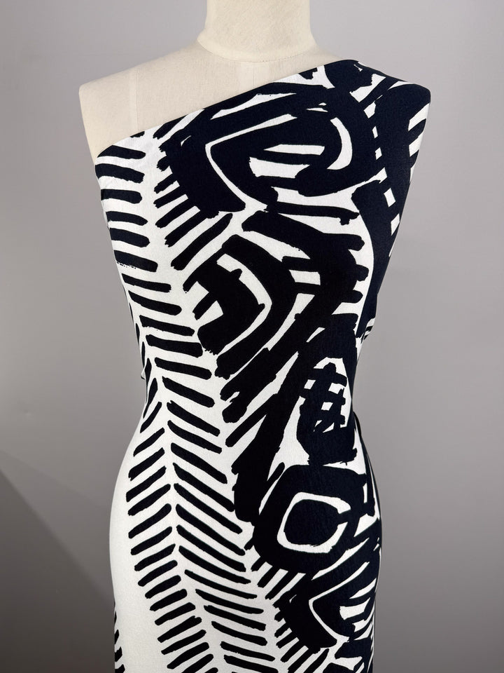Super Cheap Fabrics' Viscose Knit - Tribal Brushstroke (142cm), shown draped on a dress form, features a bold black abstract pattern on white, broad brushstroke lines and shapes, perfect for breathable summer styles like chic one-shoulder dresses.