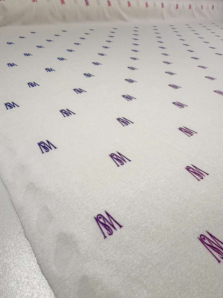 A close-up of Super Cheap Fabrics' Viscose Knit - Orchid (135cm) shows breathable white fabric with rows of blue and pink "SM" initials embroidered in a repeating pattern.
