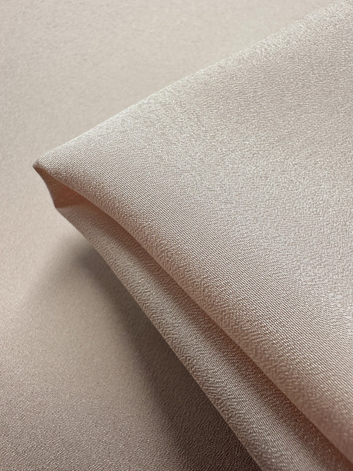 Close-up of folded mauve chalk crepe fabric from Super Cheap Fabrics, 150cm wide, with a light beige hue and subtle sheen. Smooth, soft texture enhanced by gentle lighting emphasizing its silky surface.
