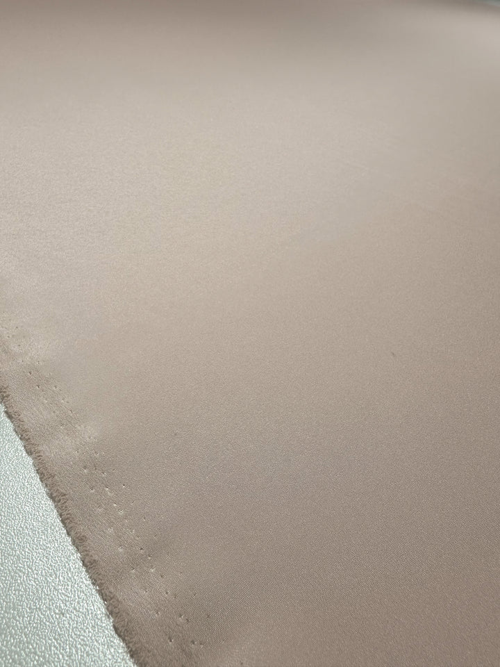 A close-up of Super Cheap Fabrics' Crepe - Mauve Chalk - 150cm features a smooth, shiny satin-like surface. The beige texture reflects light for soft highlights, and it appears flat across the surface, reminiscent of polyester crepe.