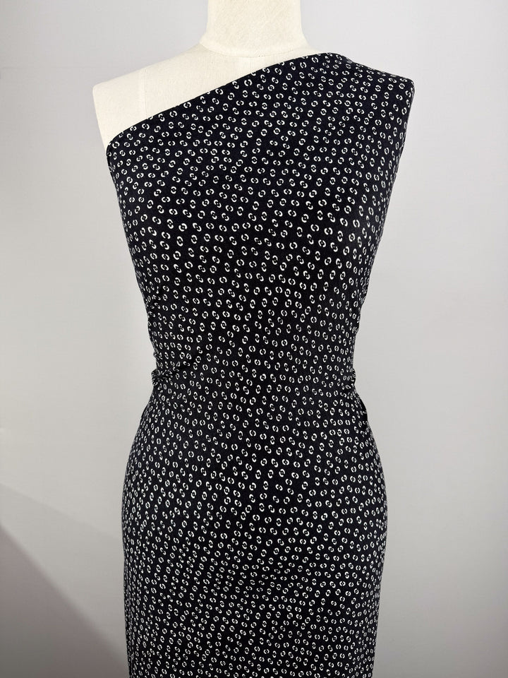 A mannequin showcases a fitted, one-shoulder dress made from Super Cheap Fabrics’ Viscose Knit - Micro Dot (145cm), featuring a black and white dotted pattern, set against a plain light gray background.