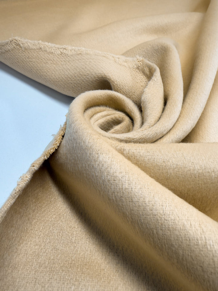 A close-up of Super Cheap Fabrics’ Wool Cashmere - Sahara Sun (150cm), extra heavy weight in light tan, shows its luxurious softness and slight fuzziness, partially rolled in a spiral with frayed edges on the left.