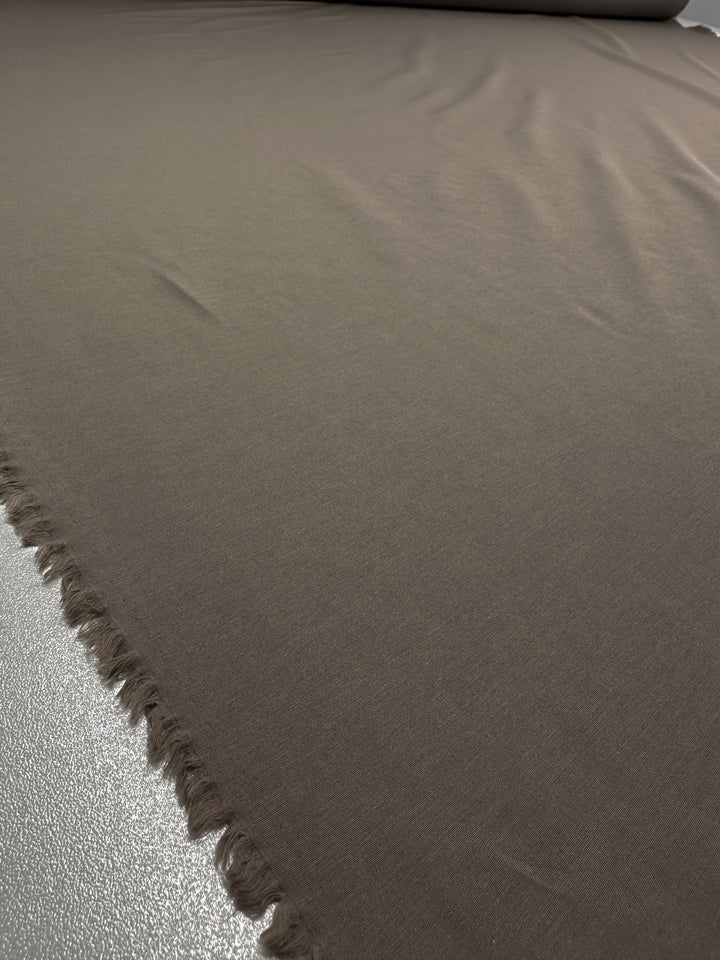 A large piece of Lightweight Bengaline - Moon Rock - 152cm by Super Cheap Fabrics is spread out on a reflective surface, with one edge slightly frayed. The smooth, lightweight fabric catches the subtle lighting, elegantly highlighting its texture.