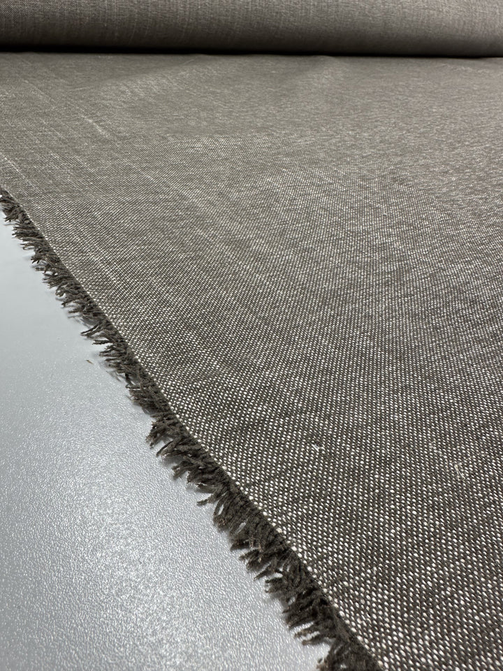 The Upholstery - Shitake – 142cm by Super Cheap Fabrics showcases a roll of grey fabric with a subtle weave and small fringed edges placed on a reflective surface. This viscose polyester linen blend is sturdy and textured, ideal for modern minimalist decor.