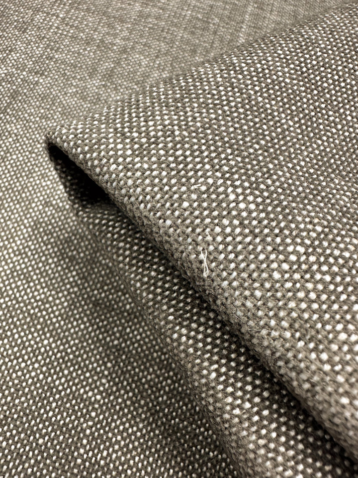 A close-up view of Super Cheap Fabrics' Upholstery - Shitake - 142cm showcases its textured grey and white woven design in a modern minimalist style with a small square pattern. A fold highlights the plain weave detail and thickness.