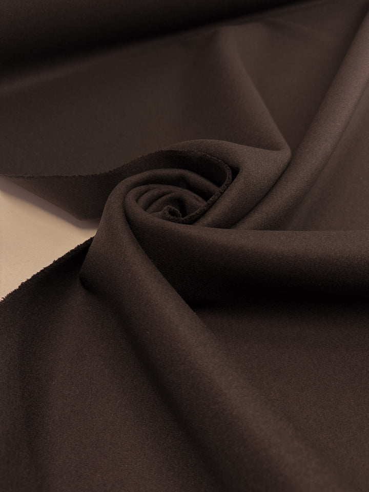 A close-up of Super Cheap Fabrics' Melton Wool - Black Coffee - 150cm, elegantly draped and rolled to showcase its smooth texture, dark brown hue, and gentle folds.