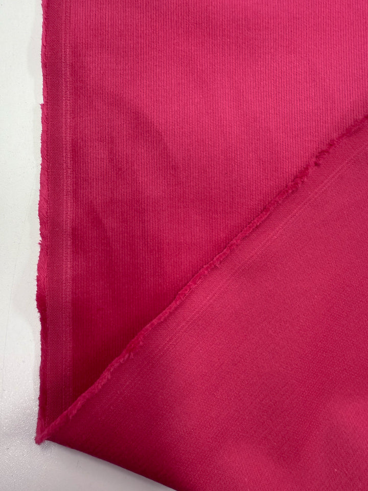 A close-up of two overlapping pieces of Super Cheap Fabrics' Pin Wale Corduroy - Love Potion (147cm), highlighting the bright pink color, textured weave, raw edges, and subtle shade variations on a white surface.