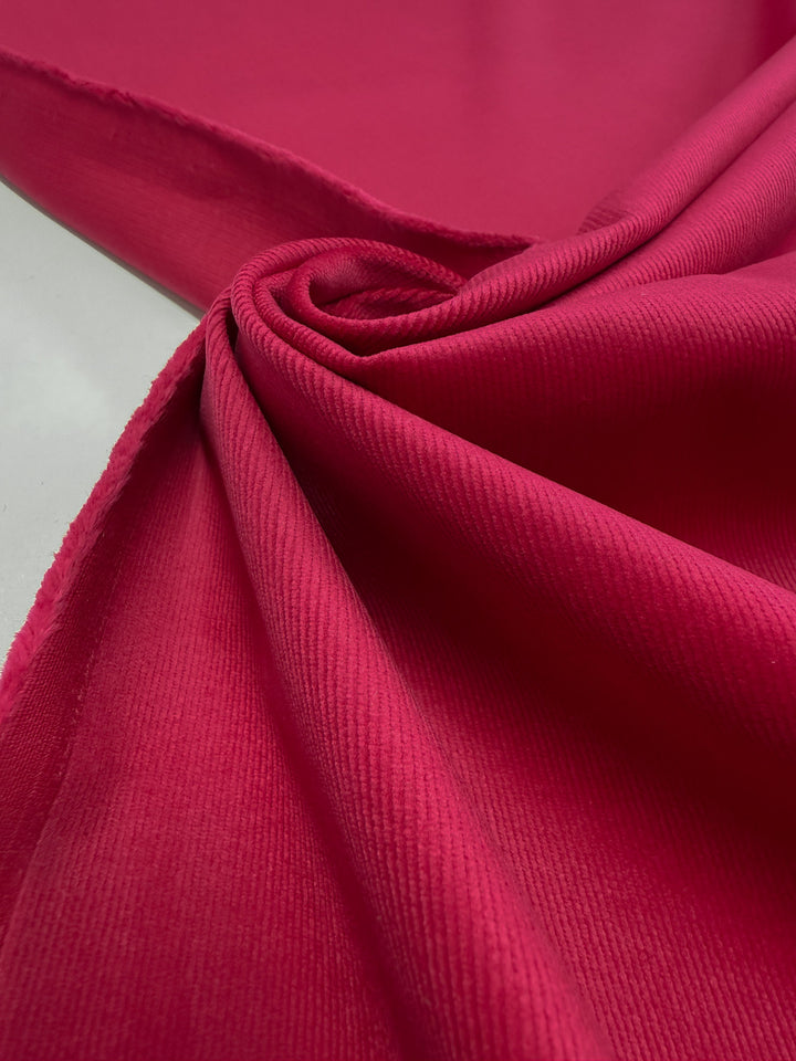 A close-up of Super Cheap Fabrics' Pin Wale Corduroy - Love Potion (147cm), highlighting its vibrant pink color, soft ridges, and visible unfinished edge on a smooth surface.
