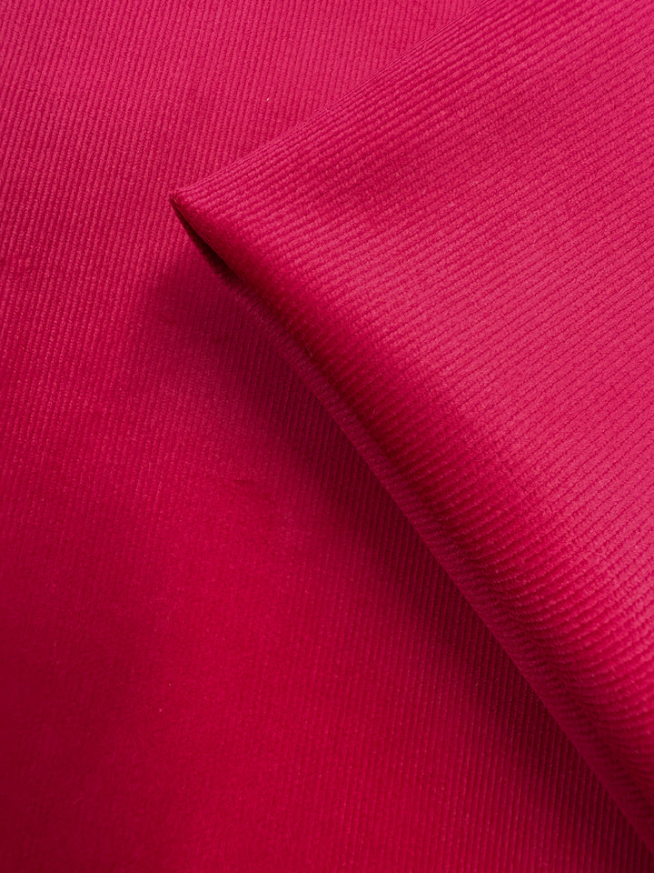 A close-up of Super Cheap Fabrics’ Pin Wale Corduroy - Love Potion (147cm), showing its bright pink color, fine ribbed texture, and smooth, medium weight cotton fabric with a neatly folded corner.