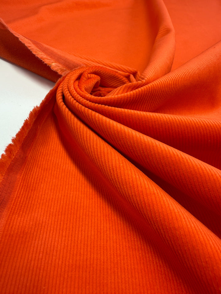 Super Cheap Fabrics' Pin Wale Corduroy - Fiesta - 142cm features soft, ribbed bright orange cotton with a velvety touch, shown gathered in a swirl to highlight its cozy texture; edges display slight fraying—ideal for warm winter wear.
