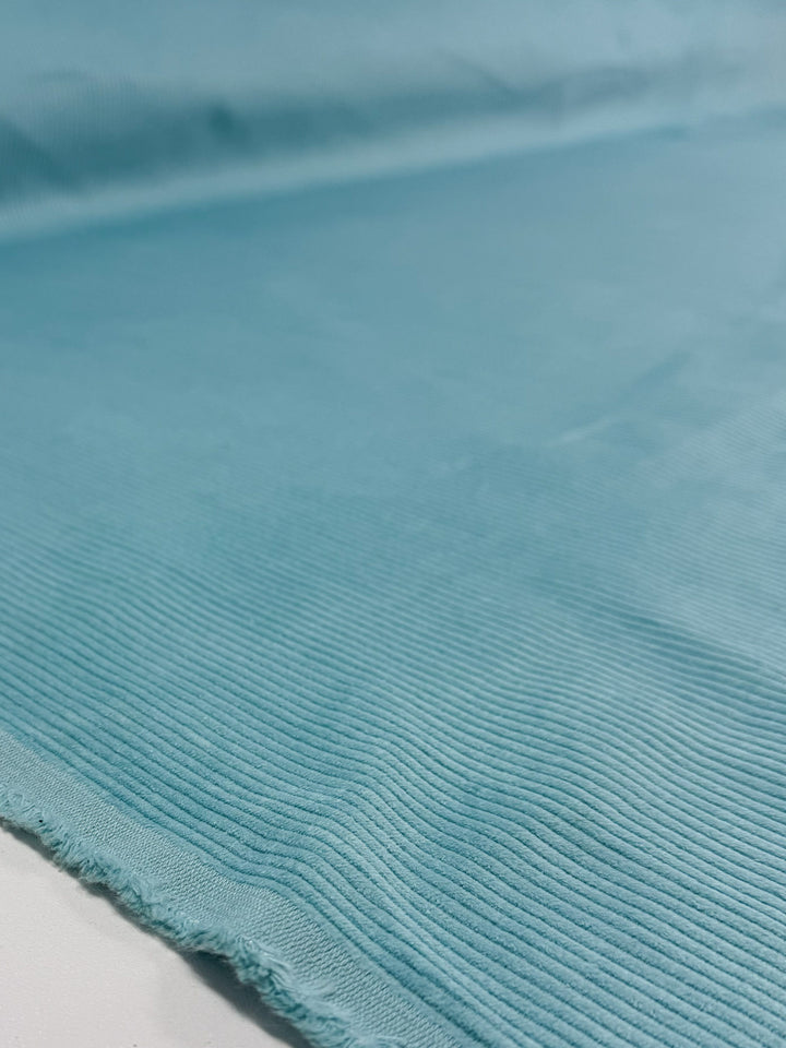 A close-up of Super Cheap Fabrics' Mid Wale Corduroy in Pastel Turquoise (145cm), highlighting its soft, textured ridges and subtle horizontal lines on flat cotton fabric with a slightly frayed edge.