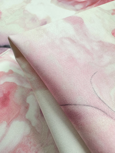 A close-up of Super Cheap Fabrics' Cotton Sateen - Mystic Garden - 125cm reveals medium weight fabric with pink and white watercolor patterns. The gently folded cotton displays soft gradients and subtle texture, offering an artistic, delicate look perfect for household décor.