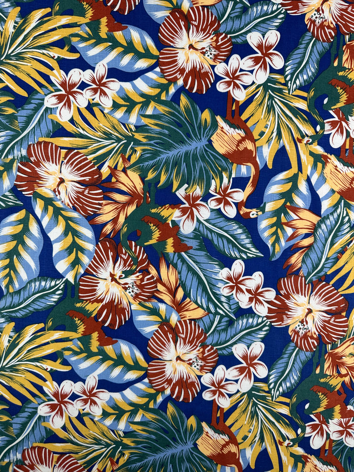 The Pure Printed Linen - Lost by Super Cheap Fabrics showcases a vibrant tropical pattern with large flowers, leaves, and flamingos on a multi-colored blue background. It features red, yellow, green, and white hues for a lightweight and exotic look. Width: 135cm.