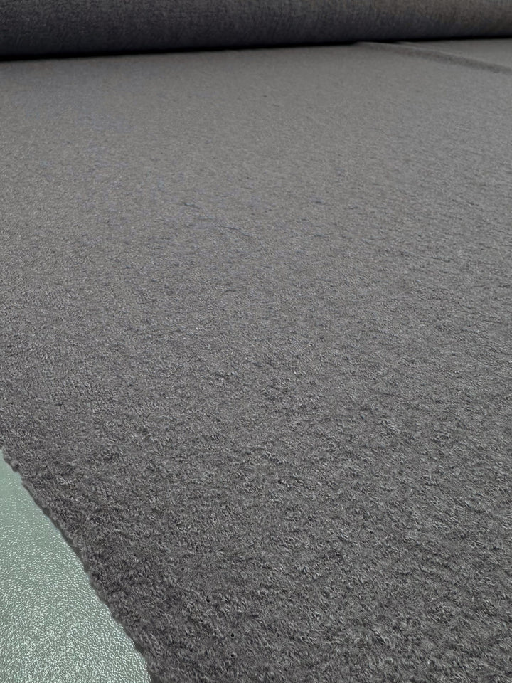 A close-up of the textured grey "Boiled Wool - Gull - 142cm" by Super Cheap Fabrics shows it partially unrolled, resembling a heavyweight wool-viscose blend. The fibrous edge of the fabric meets a greenish surface in the foreground.