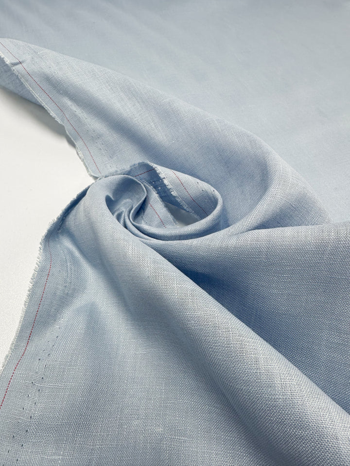 The Pure Linen - Powder Blue fabric by Super Cheap Fabrics is displayed flat with a twisted corner to showcase its texture. A subtle red stitching adds contrast, enhancing the breathable and stylishly comfortable material.